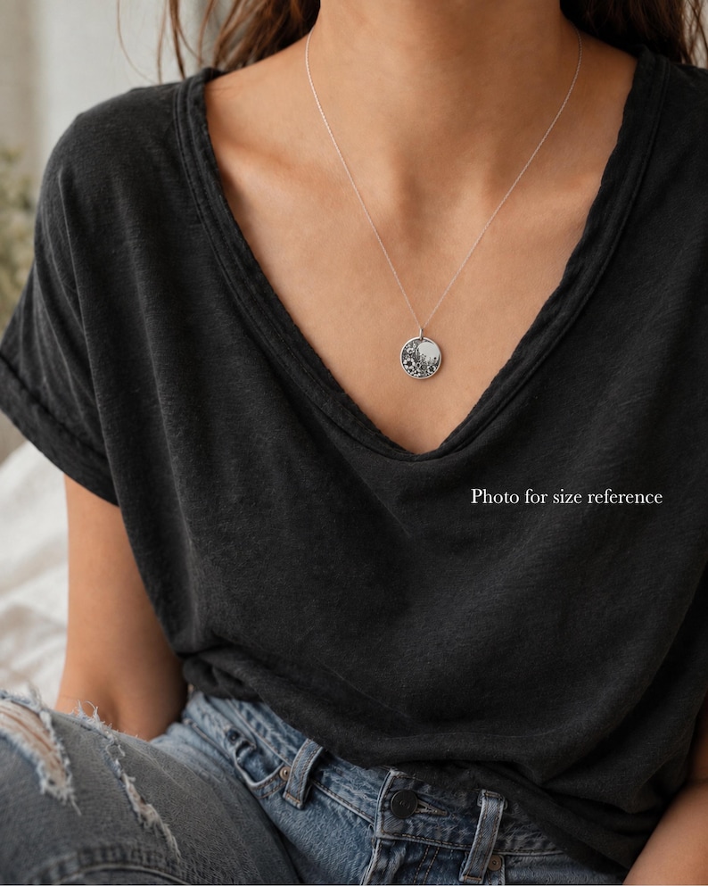 May include: A silver necklace featuring a small, round pendant with a detailed floral engraving. The pendant hangs from a delicate, thin silver chain, resting against a dark charcoal gray v-neck t-shirt and blue denim jeans.