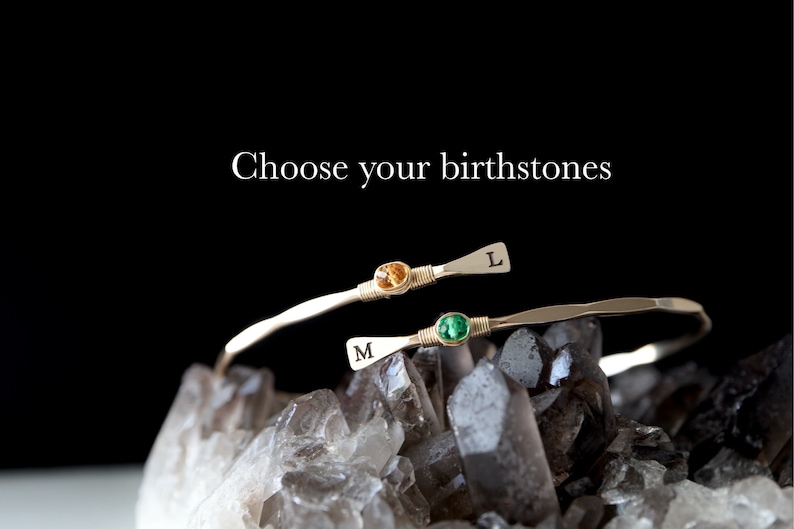 May include: Two gold bracelets with birthstones and initials. The top bracelet has an orange stone and the letter "L" engraved on it. The bottom bracelet has a green stone and the letter "M" engraved on it. The bracelets are on a bed of clear quartz crystals.