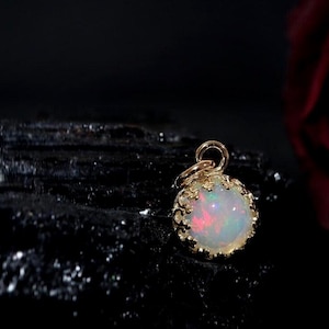 Puede incluir: Un colgante de &oacute;palo redondo engastado en un bisel de metal dorado estilo corona con una peque&ntilde;a anilla circular en la parte superior. El &oacute;palo muestra destellos iridiscentes de luz roja, verde y azul sobre una base blanca lechosa.