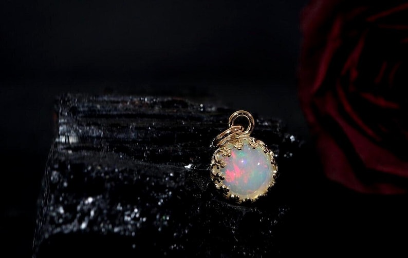 Puede incluir: Un colgante de &oacute;palo redondo engastado en un bisel de metal dorado estilo corona con una peque&ntilde;a anilla circular en la parte superior. El &oacute;palo muestra destellos iridiscentes de luz roja, verde y azul sobre una base blanca lechosa.