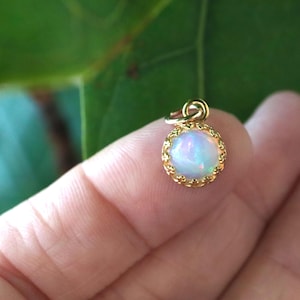 Puede incluir: Un peque&ntilde;o colgante redondo con un &oacute;palo cabuj&oacute;n que muestra destellos iridiscentes azules y rosas, engastado en un bisel de metal dorado con un borde delicado en forma de corona festoneada y una peque&ntilde;a anilla superior.