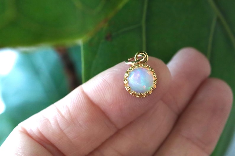 Puede incluir: Un peque&ntilde;o colgante redondo con un &oacute;palo cabuj&oacute;n que muestra destellos iridiscentes azules y rosas, engastado en un bisel de metal dorado con un borde delicado en forma de corona festoneada y una peque&ntilde;a anilla superior.