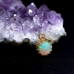 Puede incluir: Un colgante de &oacute;palo redondo engastado en un bisel dorado con forma de corona y una peque&ntilde;a anilla en la parte superior. El &oacute;palo muestra destellos iridiscentes de luz verde y naranja. El colgante est&aacute; colocado frente a un grupo de cristales de amatista.