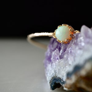 Puede incluir: Un anillo de oro con una piedra preciosa opalescente. La banda del anillo tiene un dise&ntilde;o texturizado, y la piedra preciosa est&aacute; engastada en un engaste decorativo calado. El anillo se exhibe sobre una formaci&oacute;n de cristal p&uacute;rpura.