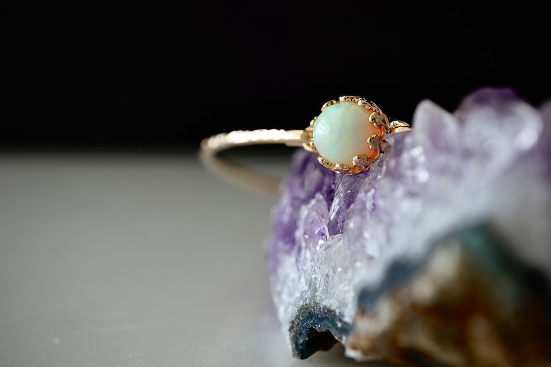 Puede incluir: Un anillo de oro con una piedra preciosa opalescente. La banda del anillo tiene un dise&ntilde;o texturizado, y la piedra preciosa est&aacute; engastada en un engaste decorativo calado. El anillo se exhibe sobre una formaci&oacute;n de cristal p&uacute;rpura.