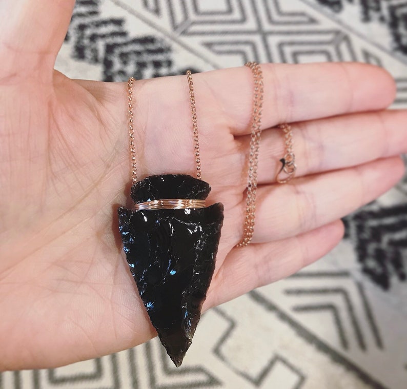 Op de afbeelding: Een zwarte obsidianen pijlpunt hanger aan een ros&eacute;gouden ketting. De pijlpunt is omwikkeld met een dunne band van ros&eacute;gouden draad.