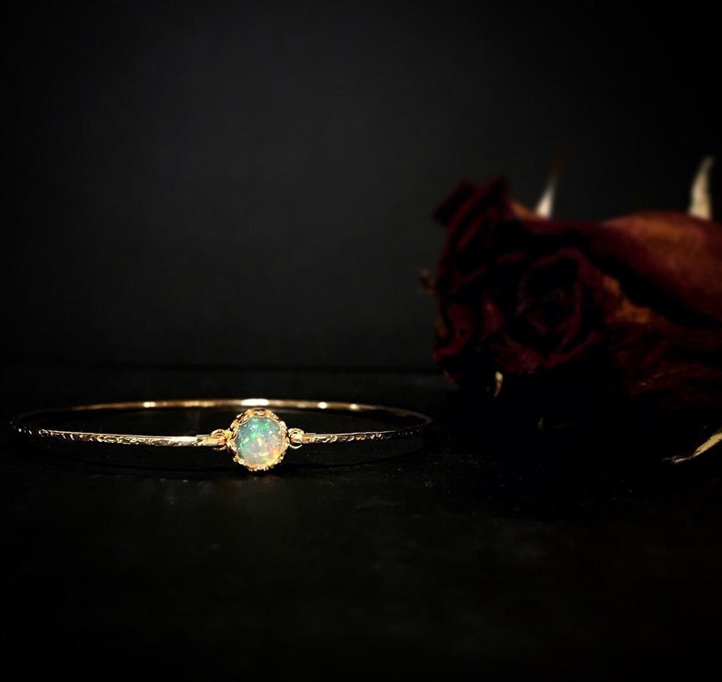 Puede incluir: Un brazalete de oro con una piedra de &oacute;palo ovalada en el centro. El &oacute;palo est&aacute; engastado en un engaste de oro decorativo. El brazalete tiene una superficie texturizada y se muestra sobre un fondo oscuro. El di&aacute;metro del brazalete es de unos 6 cm.