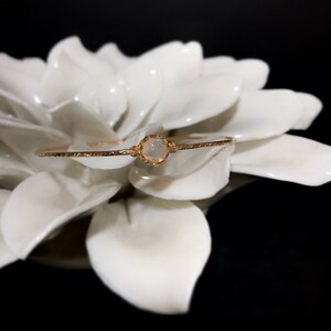 Puede incluir: Una delicada pulsera dorada con una piedra preciosa de &oacute;palo redonda central. El &oacute;palo est&aacute; engastado en un bisel decorativo, y la pulsera tiene un aspecto texturizado y martillado. La joya se exhibe sobre un fondo floral blanco.