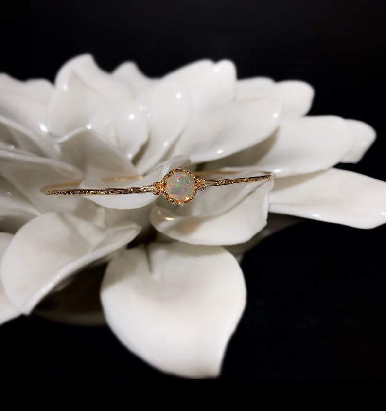 Puede incluir: Una delicada pulsera dorada con una piedra preciosa de &oacute;palo redonda central. El &oacute;palo est&aacute; engastado en un bisel decorativo, y la pulsera tiene un aspecto texturizado y martillado. La joya se exhibe sobre un fondo floral blanco.