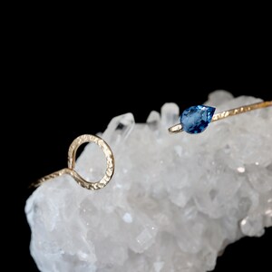 May include: A gold-toned open cuff bracelet featuring a textured, hammered metal finish. One end of the bracelet terminates in a circular loop, while the other end is accented with a single, faceted, pear-shaped blue gemstone set in metal.