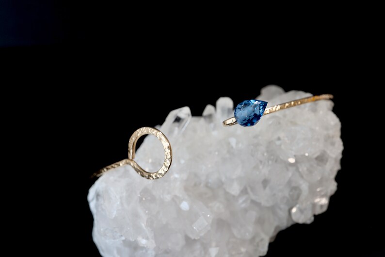 May include: A gold-toned open cuff bracelet featuring a textured, hammered metal finish. One end of the bracelet terminates in a circular loop, while the other end is accented with a single, faceted, pear-shaped blue gemstone set in metal.