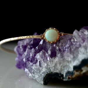 Puede incluir: Una delicada pulsera dorada con una piedra de &oacute;palo p&aacute;lido y redonda en el centro. La pulsera se coloca sobre un fondo de cristal de amatista p&uacute;rpura, creando un contraste de texturas y colores. El dise&ntilde;o de la pulsera es simple pero elegante.