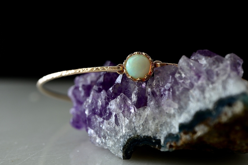 Puede incluir: Una delicada pulsera dorada con una piedra de &oacute;palo p&aacute;lido y redonda en el centro. La pulsera se coloca sobre un fondo de cristal de amatista p&uacute;rpura, creando un contraste de texturas y colores. El dise&ntilde;o de la pulsera es simple pero elegante.