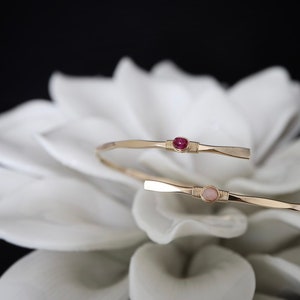May include: Two gold wire wrapped bracelets with a pink and a red gemstone. The bracelets are on a white flower.