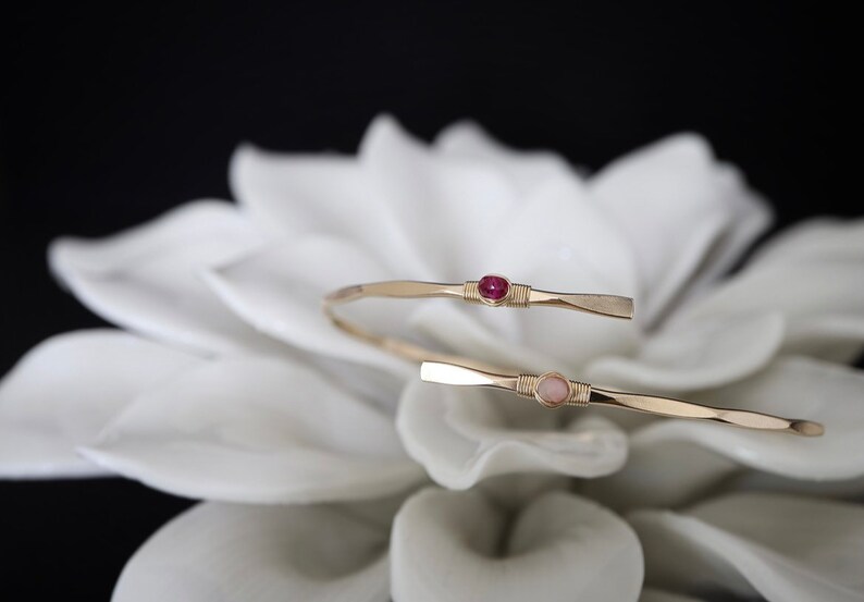 May include: Two gold wire wrapped bracelets with a pink and a red gemstone. The bracelets are on a white flower.