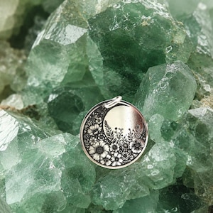 May include: A circular silver-toned pendant featuring an engraved crescent moon shape filled with detailed floral patterns including various blossoms, leaves, and stems, with a small metal jump ring attached to the top edge of the disc.