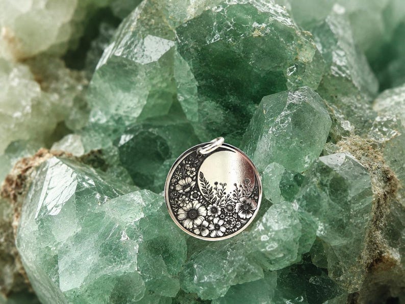 May include: A circular silver-toned pendant featuring an engraved crescent moon shape filled with detailed floral patterns including various blossoms, leaves, and stems, with a small metal jump ring attached to the top edge of the disc.
