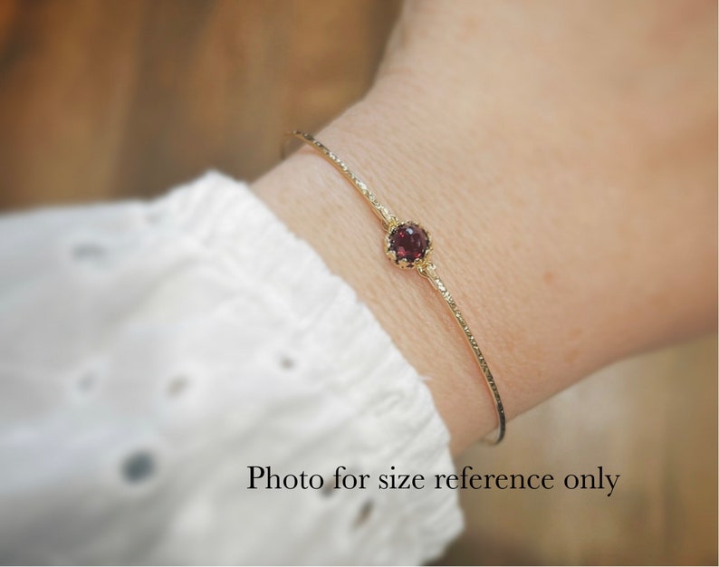 Pode incluir: Um bracelete de ouro com uma &uacute;nica pedra preciosa vermelha engastada em uma moldura de ouro. O bracelete est&aacute; no pulso de uma pessoa. Foto apenas para refer&ecirc;ncia de tamanho.