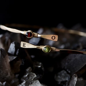 May include: Two gold bracelets with a red gemstone and a green gemstone. The bracelets are engraved with the letters "E" and "O".