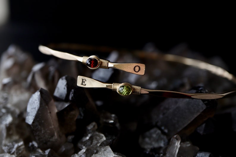 May include: Two gold bracelets with a red gemstone and a green gemstone. The bracelets are engraved with the letters "E" and "O".