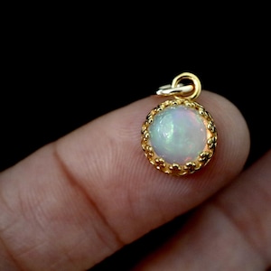 Puede incluir: Un peque&ntilde;o colgante redondo con un &oacute;palo cabuj&oacute;n que muestra destellos iridiscentes de color, engastado en un bisel de metal dorado con borde festoneado y una anilla circular en la parte superior para colgarlo de una cadena.