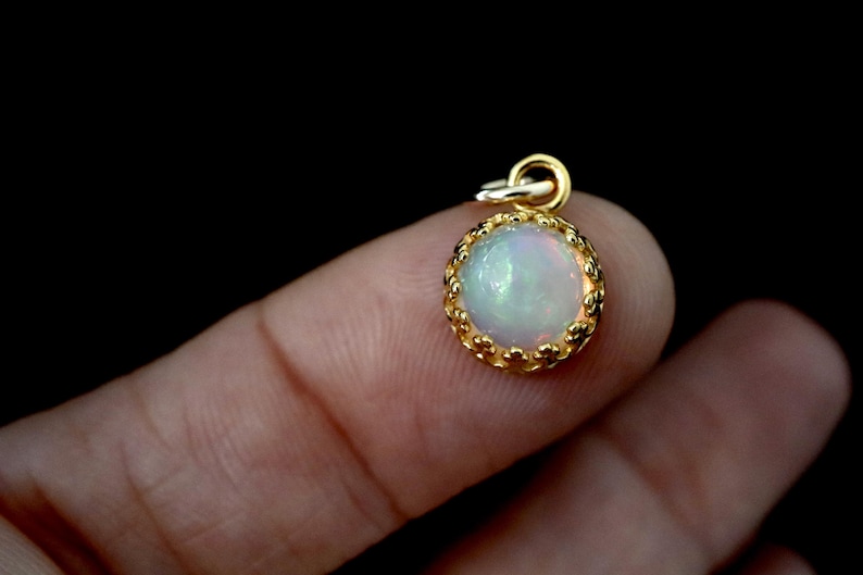 Puede incluir: Un peque&ntilde;o colgante redondo con un &oacute;palo cabuj&oacute;n que muestra destellos iridiscentes de color, engastado en un bisel de metal dorado con borde festoneado y una anilla circular en la parte superior para colgarlo de una cadena.