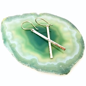 May include: A pair of silver hammered bar earrings with a delicate wire hook closure. The earrings are displayed on a green and white agate slice.