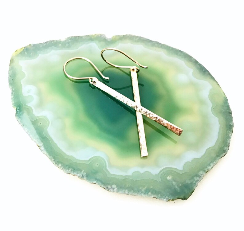 May include: A pair of silver hammered bar earrings with a delicate wire hook closure. The earrings are displayed on a green and white agate slice.