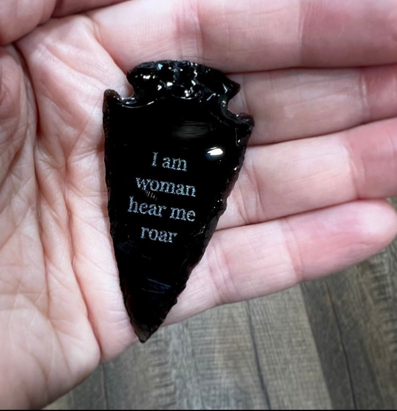 Op de afbeelding: Een zwarte obsidianen pijlpunt met de woorden "I am woman hear me roar" erop gegraveerd.