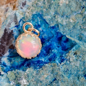 Puede incluir: Un colgante de &oacute;palo redondo con corte cabuj&oacute;n engastado en un bisel de metal dorado con borde festoneado. El &oacute;palo muestra una base blanca lechosa con destellos iridiscentes de color rosa y naranja. Una peque&ntilde;a anilla dorada est&aacute; sujeta en la parte superior.