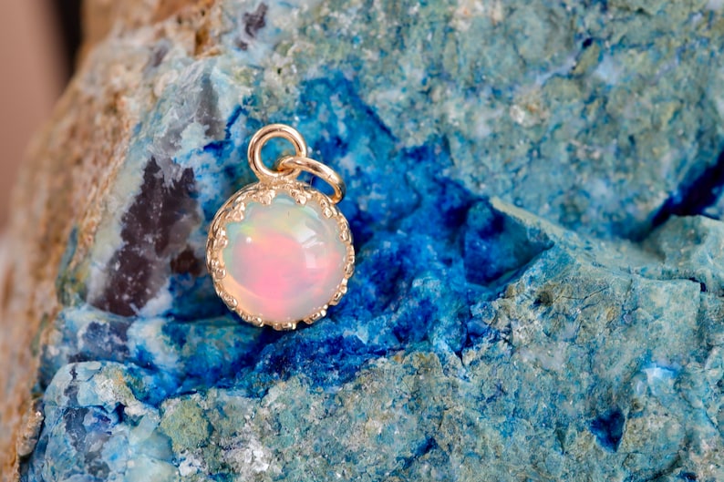 Puede incluir: Un colgante de &oacute;palo redondo con corte cabuj&oacute;n engastado en un bisel de metal dorado con borde festoneado. El &oacute;palo muestra una base blanca lechosa con destellos iridiscentes de color rosa y naranja. Una peque&ntilde;a anilla dorada est&aacute; sujeta en la parte superior.