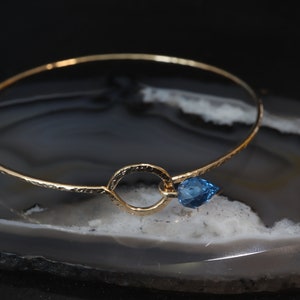 May include: A thin gold bangle bracelet featuring a textured, hammered surface. The bracelet includes a central circular loop element that holds a single, small, faceted, pear-shaped blue gemstone charm attached to the side of the ring.