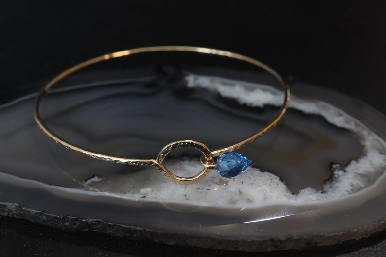 May include: A thin gold bangle bracelet featuring a textured, hammered surface. The bracelet includes a central circular loop element that holds a single, small, faceted, pear-shaped blue gemstone charm attached to the side of the ring.