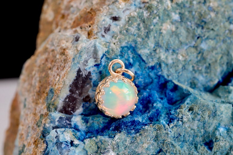 Puede incluir: Un colgante redondo con una piedra de &oacute;palo iridiscente lisa engastada en un bisel de metal dorado con un borde festoneado decorativo y una peque&ntilde;a anilla circular en la parte superior para colgar de una cadena.