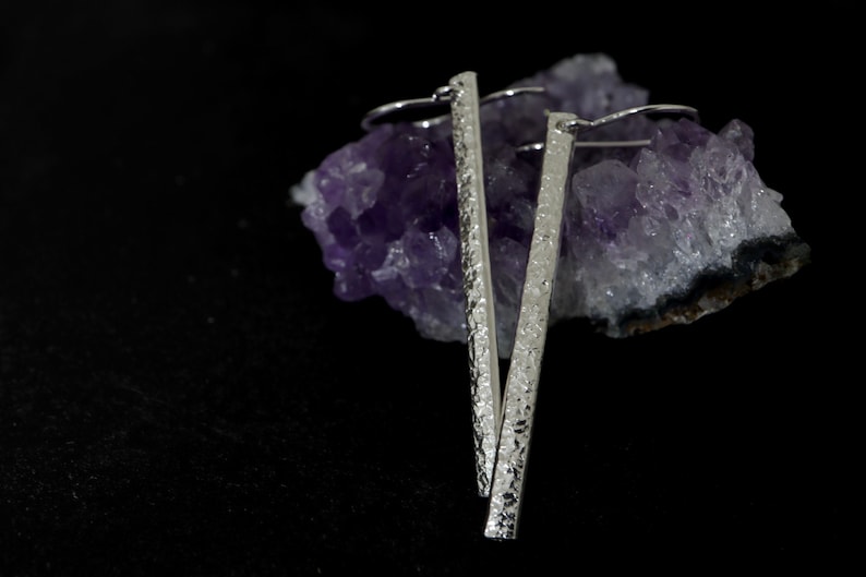 May include: A pair of silver earrings with a textured, hammered finish. The earrings are long and slender, and they are hanging from a cluster of amethyst crystals.