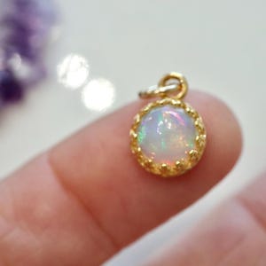 Puede incluir: Un peque&ntilde;o colgante redondo con un &oacute;palo cabuj&oacute;n que muestra destellos iridiscentes, engastado en un bisel de metal dorado con borde festoneado y una peque&ntilde;a anilla en la parte superior para colgarlo de una cadena.