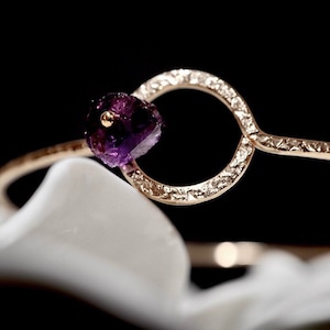 May include: A gold bangle bracelet with a textured finish and a single purple gemstone. The bangle is open and has a circular design.