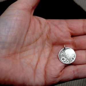 May include: A circular silver pendant featuring an engraved design of a crescent moon and a cluster of various small flowers. The pendant has a small metal loop at the top for attachment to a chain or cord, showcasing a detailed floral pattern.