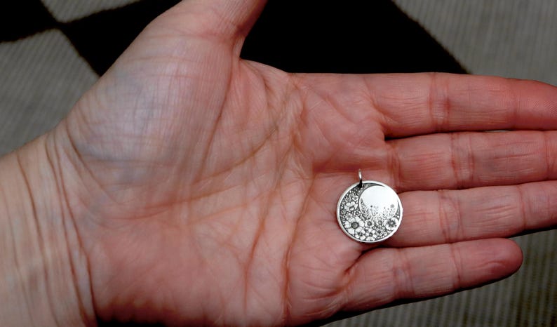 May include: A circular silver pendant featuring an engraved design of a crescent moon and a cluster of various small flowers. The pendant has a small metal loop at the top for attachment to a chain or cord, showcasing a detailed floral pattern.