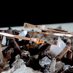 May include: Two gold wire-wrapped bracelets with gemstone accents. The top bracelet features a clear gemstone and the letter "A" engraved on the end. The bottom bracelet features a yellow gemstone and the letter "R" engraved on the end. The bracelets are displayed on a bed of clear quartz crystals.