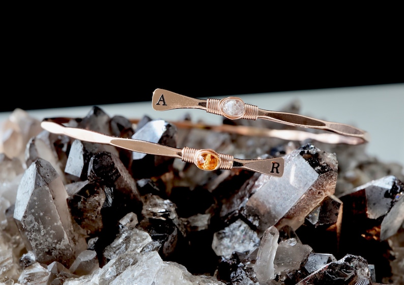 May include: Two gold wire-wrapped bracelets with gemstone accents. The top bracelet features a clear gemstone and the letter "A" engraved on the end. The bottom bracelet features a yellow gemstone and the letter "R" engraved on the end. The bracelets are displayed on a bed of clear quartz crystals.