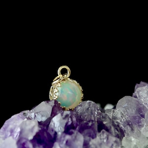 Puede incluir: Un colgante de &oacute;palo iridiscente de forma redonda engastado en un marco de metal dorado estilo corona con una peque&ntilde;a anilla circular en la parte superior, sobre un grupo de cristales de amatista violeta oscuro con fondo negro.