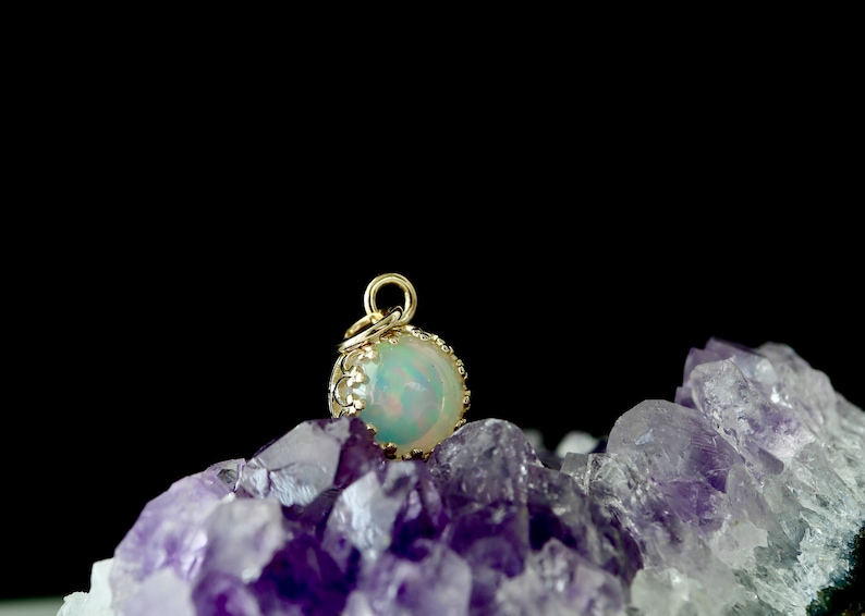 Puede incluir: Un colgante de &oacute;palo iridiscente de forma redonda engastado en un marco de metal dorado estilo corona con una peque&ntilde;a anilla circular en la parte superior, sobre un grupo de cristales de amatista violeta oscuro con fondo negro.