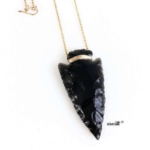 Op de afbeelding: Een ketting met een hanger in de vorm van een zwarte obsidianen pijlpunt, een gouden ketting en een gouden draadwikkeling om de pijlpunt. De pijlpunt is een natuursteen en heeft een unieke vorm.