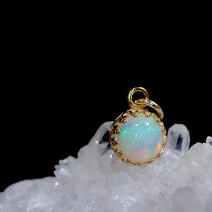 Puede incluir: Un cabuj&oacute;n de &oacute;palo redondo e iridiscente engastado en un colgante de metal dorado con estilo de corona. La piedra preciosa muestra destellos de luz verde, azul y naranja. El colgante tiene una peque&ntilde;a anilla circular arriba.