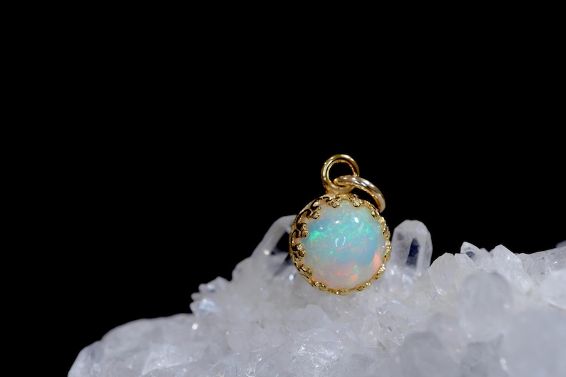 Puede incluir: Un cabuj&oacute;n de &oacute;palo redondo e iridiscente engastado en un colgante de metal dorado con estilo de corona. La piedra preciosa muestra destellos de luz verde, azul y naranja. El colgante tiene una peque&ntilde;a anilla circular arriba.