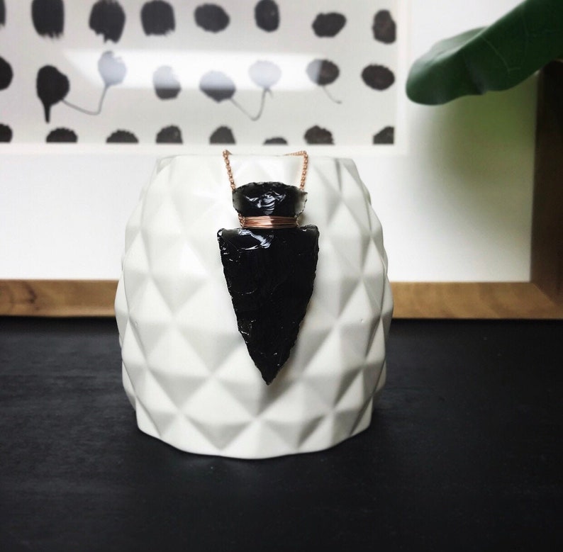 Op de afbeelding: Een zwarte obsidianen pijlpunt hangerketting met een koperen draadwikkeling. De ketting wordt getoond op een witte keramische vaas met een textuur van een diamantpatroon.