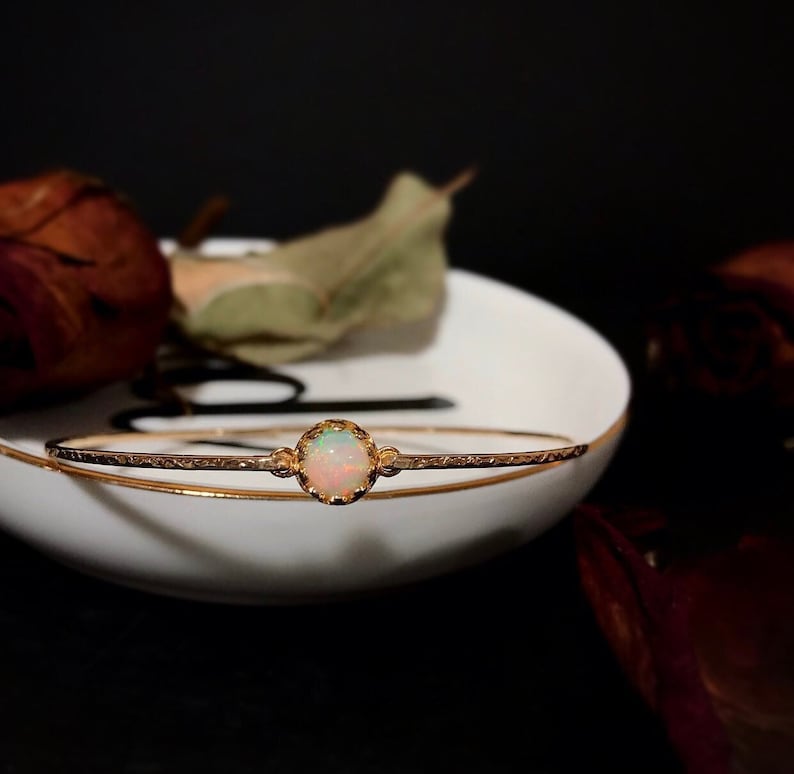 Puede incluir: Un delicado brazalete dorado con una piedra preciosa de &oacute;palo en el centro. El &oacute;palo es redondo y est&aacute; engastado en un bisel decorativo. El brazalete descansa sobre un plato blanco con borde dorado, sobre un fondo oscuro.