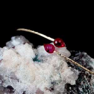May include: A delicate gold bangle bracelet with two red gemstone accents. The bracelet is textured and has a hammered finish.