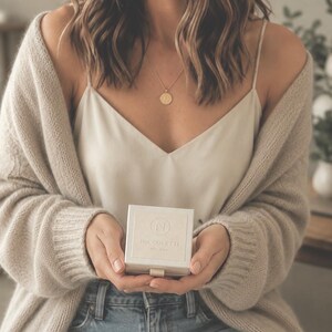 May include: A square, off-white jewelry box with a small pull tab, featuring the embossed letter N inside a circle and the text NICCOLETTE EST. 2018. The box is held in place, accompanied by a gold-toned circular pendant necklace and a beige cardigan.
